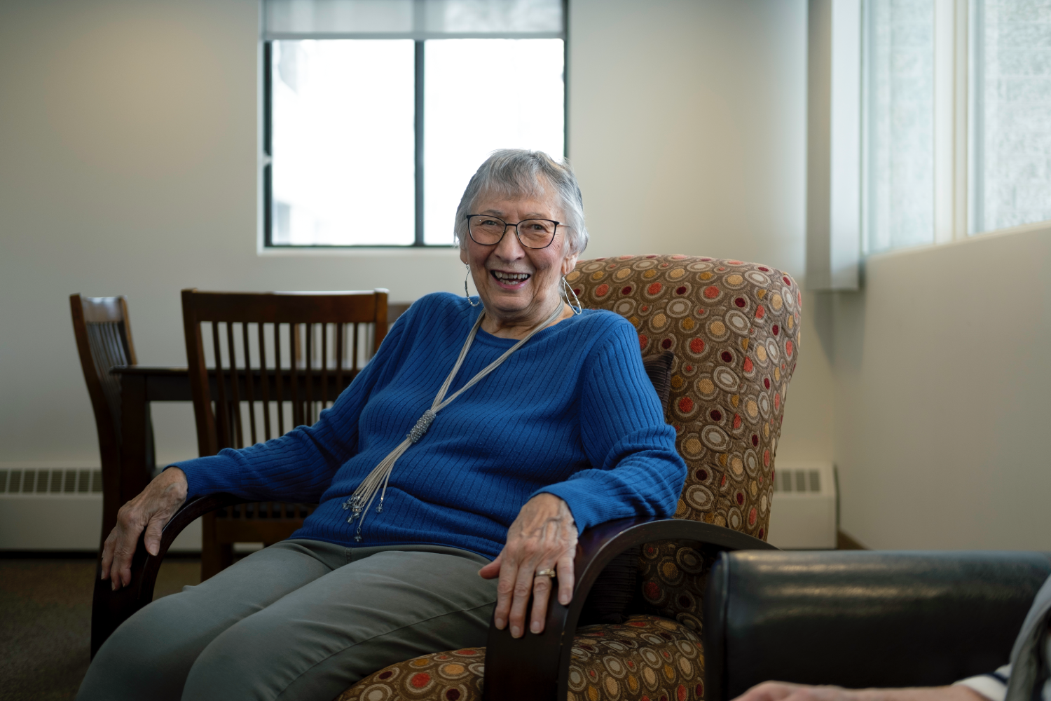 Smiling senior woman in a blue sweater sits in a patterned armchair in a light-filled room. Smiling senior woman in a blue sweater sits in a patterned armchair in a light-filled room.